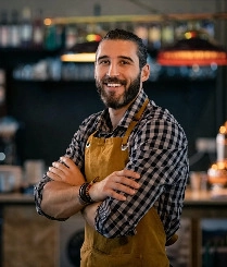 A smiling man wearing an apron stands confidently in front of a well-stocked bar.